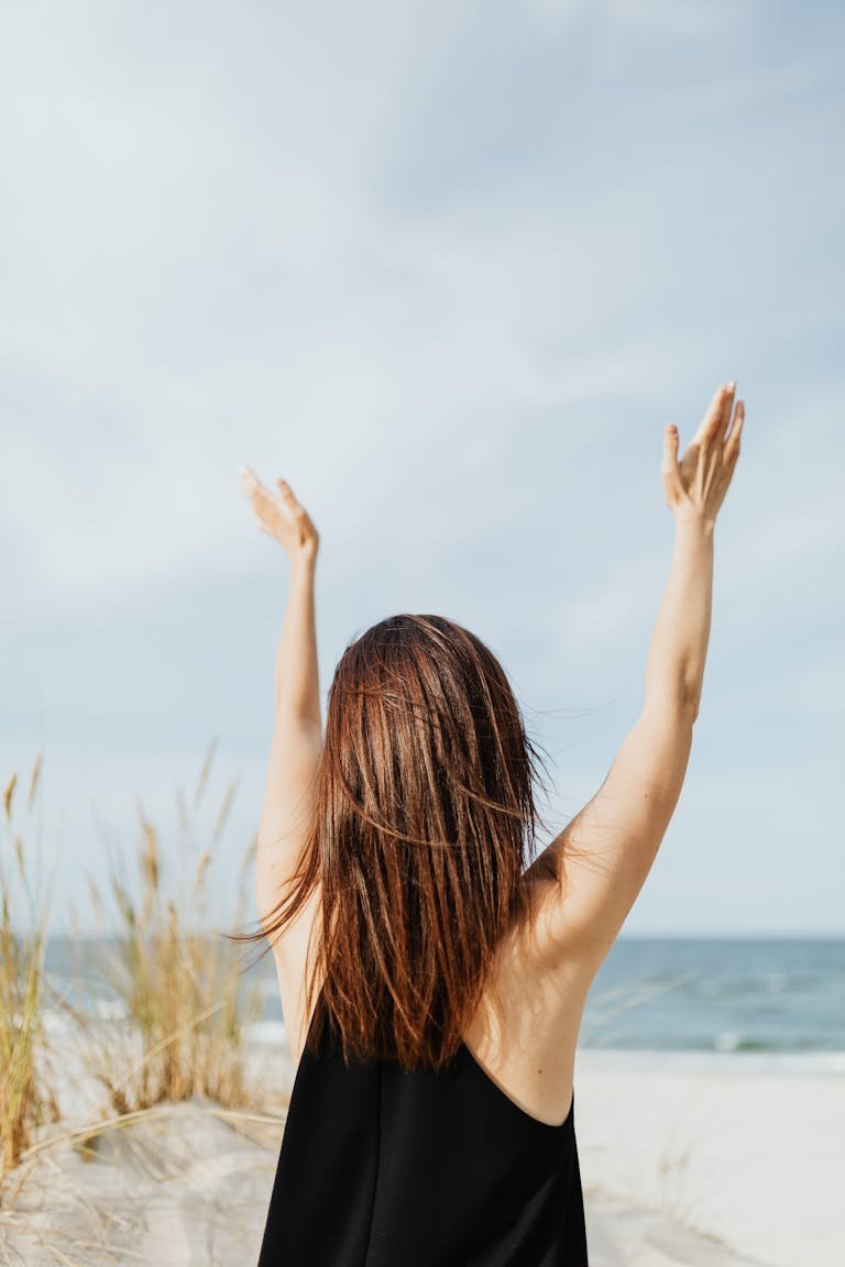 A woman joyfully raises her arms on a sandy beach with a blue sky backdrop.
